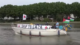 L'image montre un bateau naviguant sur une étendue d'eau. Sur le bateau, il y a des personnes présentes, et une banderole est accrochée, indiquant "SHIP TO GAZA". Le bateau arbore également plusieurs drapeaux, dont ceux de la Palestine et de la Suède, ainsi que d'autres drapeaux qui pourraient représenter différentes organisations ou pays. En arrière-plan, des arbres et des bâtiments sont visibles, suggérant un environnement urbain proche d'une rivière ou d'un port.
