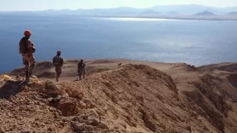 L'image montre un paysage désertique avec des militaires en patrouille sur une crête surplombant une étendue d'eau. Le terrain est aride et rocheux, avec des montagnes en arrière-plan. Les soldats portent des uniformes et des casques, et semblent observer l'horizon, avec un ciel clair et ensoleillé qui ajoute à l'atmosphère de la scène.