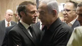 L'image montre deux hommes en discussion proche l'un de l'autre. L'un est en costume noir et le second porte une chemise noire. Ils semblent engageants dans une conversation sérieuse, entourés d'autres personnes en arrière-plan. L'ambiance générale évoque une rencontre formelle ou un événement important.