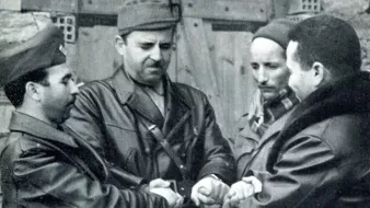 L'image montre quatre hommes serrant les mains les uns des autres dans une posture de camaraderie ou de négociation. Ils portent des tenues militaires ou de combat, et l'environnement semble être extérieur, avec des murs en pierre en arrière-plan. L'expression de leurs visages peut suggérer la détermination ou la concentration, et l'interaction semble exprimer un moment significatif.