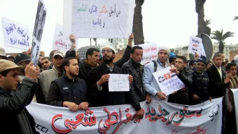 L'image montre un groupe de personnes lors d'une manifestation. Elles tiennent des pancartes et une grande banderole sur laquelle est inscrit en arabe et en français "Pour une constitution démocratique". Les manifestants semblent unis dans leur revendication, et l'environnement montre qu'ils se rassemblent probablement dans une ville, avec des palmiers en arrière-plan. L'atmosphère semble engagée et déterminée.