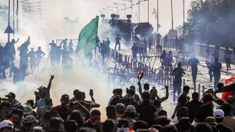 L'image montre une scène de tumultes ou de manifestations. On y voit un grand groupe de personnes rassemblées, brandissant des drapeaux et levant les bras. Il y a des barrages en bois et de la fumée, probablement causée par des gaz lacrymogènes. À l'arrière-plan, des véhicules blindés sont visibles, renforçant l'idée d'une situation tendue. L'atmosphère est chargée et chaotique, avec des manifestants qui semblent déterminés et en mouvement.