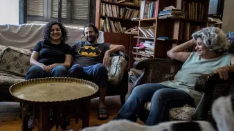 L'image montre un groupe de trois personnes assises dans un salon. Il y a deux femmes et un homme. La première femme, à gauche, sourit et porte un t-shirt noir. L'homme, au centre, porte un t-shirt avec le logo de Batman. La deuxième femme, à droite, a les cheveux bouclés et semble détendue. Ils sont assis sur des canapés, entourés d'étagères remplies de livres et d'objets décoratifs. Une ambiance conviviale et décontractée se dégage de la scène.