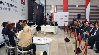 L'image montre un panel de discussion ou une session d'audition, avec plusieurs personnes assises face à une table à l'avant. Au fond, il y a une bannière affichant des informations sur l'événement, datée des 16 et 17 décembre 2016. Les participants semblent engagés dans la discussion, avec une variété de personnes, y compris des femmes portant des foulards. La scène est bien organisée, avec des cordons de sécurité délimitant l'espace où se trouvent le public et les intervenants.