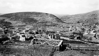 L'image montre un paysage rural en noir et blanc, où l'on peut apercevoir des ruines de structures en pierre, témoignant d'une ancienne habitation. Le terrain est vallonné, avec des collines en arrière-plan et une végétation clairsemée. Certaines zones semblent cultivées ou utilisées pour l'élevage, tandis que d'autres présentent des traces de dégradation ou d'abandon. L'atmosphère générale est paisible, mais elle évoque également une histoire de déclin ou de dévastation.