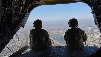 L'image montre deux membres d'équipage militaires assis à l'arrière d'un hélicoptère, regardant à l'extérieur. Ils portent des uniformes de camouflage et des casques. À travers l'ouverture de l'appareil, on peut apercevoir un paysage urbain et un plan d'eau en contrebas, sous un ciel clair. La vue offre un aperçu de la ville avec des routes et des bâtiments visibles.