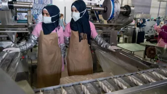 L'image montre deux femmes travaillant dans un environnement industriel. Elles portent des hijabs et des masques de protection. Elles sont habillées de vêtements de travail, avec des blouses et des tabliers. À l'arrière-plan, on peut voir des machines et d'autres personnes également en train de travailler, ce qui indique qu'il s'agit probablement d'une usine ou d'un atelier de production. L'atmosphère semble axée sur le travail et la production.