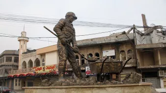L'image montre une statue monumentale représentant un travailleur en train de creuser, symbolisant probablement la reconstruction ou le labeur dans un contexte difficile. En arrière-plan, on distingue des bâtiments partiellement détruits et des infrastructures urbaines. La scène évoque une atmosphère de résilience et de reconstruction dans une zone touchée par des conflits. La statue est installée sur un socle en pierre, ajoutant à son importance et à son impact visuel.