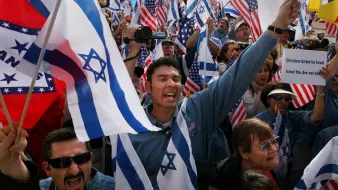 L'image montre une manifestation avec des participants brandissant des drapeaux israéliens et américains. Les gens semblent très engagés et animés, certains lèvent les bras en l'air et expriment leur enthousiasme, tandis que d'autres portent des expressions de concentration ou de soutien. Il y a un mélange de différentes personnes dans la foule, suggérant une diversité au sein de ce rassemblement.