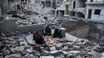 L'image montre un groupe de personnes assises sur un tapis dans un environnement dévasté. Autour d'eux, on peut voir des débris de bâtiments effondrés et des structures endommagées. Les personnes semblent concentrées sur une activité, malgré la destruction qui les entoure. L'atmosphère évoque la résilience et la vie qui continue malgré les difficultés.
