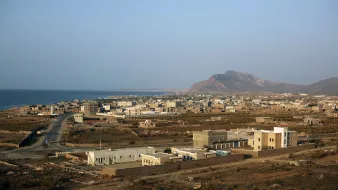 L'image montre un paysage côtier avec une vue sur une petite ville située près de la mer. On peut apercevoir des bâtiments de différentes tailles, certains modernes et d'autres plus traditionnels, dispersés sur une étendue de terrain. À l'arrière-plan, on distingue une montagne dont les contours se dessinent sous un ciel clair. La route serpente à travers la ville, menant à la mer. L'ensemble dégage une atmosphère paisible et semi-aride, typique des régions côtières.