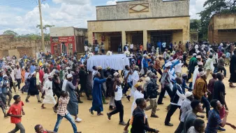L'image montre une grande foule de personnes marchant dans une rue, probablement lors d'un événement ou d'une procession. Au centre, il y a un chariot couvert d'un drap blanc, et les gens semblent marcher avec une certaine ferveur. On peut apercevoir différents styles vestimentaires, suggérant une diversité parmi les participants. En arrière-plan, des bâtiments sont visibles, ainsi qu'un ciel partiellement nuageux. L'atmosphère semble collective et déterminée.