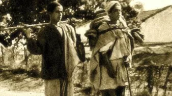 L'image montre deux personnes marchant sur un chemin. L'une d'elles est un jeune garçon, tandis que l'autre est une femme plus âgée. Les deux portent des charges sur leurs épaules, indiquant qu'ils transportent des provisions ou des biens. Le décor semble rural, avec quelques arbres en arrière-plan, et l'atmosphère générale donne un aperçu de la vie quotidienne. Le style de vêtements et le traitement de la photo évoquent une époque passée.