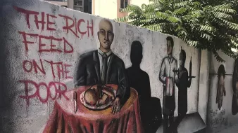 L'image montre une fresque murale colorée sur un mur. Au premier plan, un homme en costume est assis à une table couverte d'une nappe rouge, avec un plat à manger devant lui. À côté, on peut voir une ombre d'un serveur, suggérant un contexte de service alimentaire. Sur le mur, il est écrit "THE RICH FEED ON THE POOR", ce qui évoque un message critique sur les inégalités sociales. Des éléments de l'arrière-plan, comme des bâtiments et de la verdure, ajoutent à l'environnement urbain.