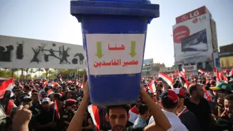 L'image montre une grande manifestation avec une foule rassemblée. Au premier plan, un homme porte un grand seau bleu sur sa tête, qui affiche des inscriptions en arabe. Derrière lui, on peut voir de nombreux drapeaux, probablement ceux de l'Égypte, et des affiches graphiques sur un mur. La scène dégage une atmosphère de protestation, avec des gens mobilisés pour une cause.