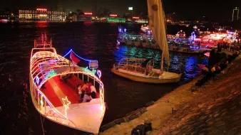 L'image montre un paysage nocturne animé le long d'une rivière. Plusieurs bateaux décorés de lumières colorées sont amarrés, créant une atmosphère festive. On peut voir des gens sur les bateaux et le long de la promenade, profitant de l'ambiance. En arrière-plan, des bâtiments illuminés ajoutent à la scène, avec des reflets dans l'eau. L'ensemble évoque une belle soirée en bord de rivière.