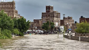 L'image montre une scène de inondation dans une ville, probablement au Yémen. On peut voir des bâtiments traditionnels en pierre avec des arches et des fenêtres ornementales. Les niveaux d'eau sont élevés, couvrant une grande partie de la rue, ce qui semble indiquer des pluies abondantes. Des gens se rassemblent sur le bord, observant la situation. Le ciel est nuageux, ajoutant à l'atmosphère dramatique de la scène. Les arbres sont verdoyants, indiquant une nature environnante peu affectée par la montée des eaux.