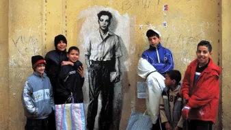L'image montre un groupe de jeunes garçons rassemblés devant un mur de couleur jaune. Sur ce mur se trouve un dessin noir et blanc représentant un homme faisant face à la caméra. Les garçons, qui portent des vêtements variés et colorés, affichent des expressions diverses, allant du sourire à la curiosité. L'ensemble de la scène évoque un moment de camaraderie et de jeu dans un environnement urbain.