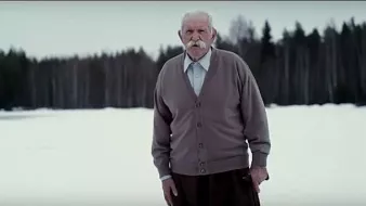 L'image montre un homme âgé se tenant debout sur un terrain enneigé. Il porte un pull en cardigan gris et un pantalon, et il a une moustache bien fournie. En arrière-plan, on peut voir une forêt de conifères qui ajoute une ambiance tranquille et froide à la scène. L'homme semble contemplatif, regardant droit devant lui dans un paysage hivernal.