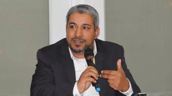 L'image montre un homme assis à une table, tenant un microphone. Il semble engager une discussion ou faire une présentation. Il porte un costume sombre avec une chemise claire et a des cheveux grisonnants. L'environnement autour de lui semble être celui d'une conférence ou d'un événement formel, avec une lumière claire et des fenêtres en arrière-plan.