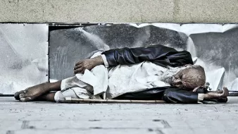 L'image montre un homme âgé allongé sur le sol, semblant endormi. Il porte des vêtements usés et semble être dans une position de repos sur un trottoir. Son visage est marqué par le temps et il est entouré d'un environnement urbain. L'atmosphère évoque une certaine mélancolie et une réflexion sur la vie des personnes sans abri.