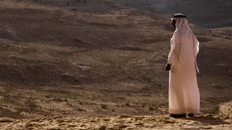 L'image montre une personne vêtue d'une tenue traditionnelle, probablement un kandora ou un dishdasha, se tenant debout sur un terrain désertique. En arrière-plan, on aperçoit un paysage aride avec des collines et des formations rocheuses. La lumière est douce, ce qui donne une ambiance calme et contemplative à la scène. La personne semble regarder au loin, peut-être en train de réfléchir ou d'admirer la vue panoramique du désert.