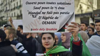 L'image montre une manifestation, où une jeune femme tient une pancarte. La pancarte indique qu'elle est ouverte à toutes solutions, sauf pour le 4ème mandat, affirmant que le mandat ne fait pas partie de l'Algérie de demain. La femme est entourée d'autres manifestants et porte un drapeau. L'atmosphère semble engagée et déterminée, reflétant des revendications politiques.