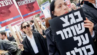 L'image montre une manifestation avec des participants portant des vêtements sombres, probablement des avocats, et tenant des pancartes. L'un des manifestants, au premier plan, tient une grande affiche noire avec du texte écrit en hébreu. L'atmosphère semble être sérieuse et engagée, avec des expressions résolues sur les visages des manifestants. En arrière-plan, d'autres personnes tiennent également des pancartes, ajoutant à l'ambiance de protestation.