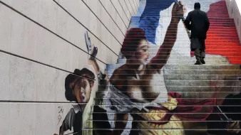 L'image montre une fresque peinte sur des marches d'escalier. On y voit une scène dynamique, représentant une femme aux vêtements colorés, brandissant une arme et symbolisant la liberté. À ses côtés, un jeune garçon semble se joindre à elle, tenant un pistolet. En arrière-plan, on remarque des teintes qui évoquent le drapeau français, avec des nuances de bleu, blanc et rouge. Une personne en noir monte les marches, ajoutant une dimension de mouvement à l'œuvre. Cette fresque combine art et engagement, reflétant des thèmes de révolution et de lutte.