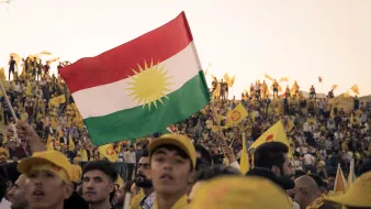 L'image montre une foule nombreuse, probablement lors d'un événement ou d'une célébration. Au premier plan, une personne brandit un drapeau kurde qui présente des couleurs vives avec un soleil stylisé. La plupart des membres de la foule portent des casquettes ou des vêtements jaunes, ajoutant à l'ambiance festive. L'arrière-plan est rempli de personnes, toutes engagées et enthousiastes, ce qui suggère une forte mobilisation et un sentiment de communauté.