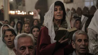 L'image montre un groupe de personnes rassemblées dans un lieu de culte. Au premier plan, une femme portant un vêtement rouge et un voile lit un livre, probablement un texte religieux. En arrière-plan, d'autres personnes, dont certaines semblent chanter ou prier, sont visibles. L'atmosphère semble solennelle, éclairée par des bougies qui créent une ambiance chaleureuse. Les visages des participants expriment une concentration et une dévotion.