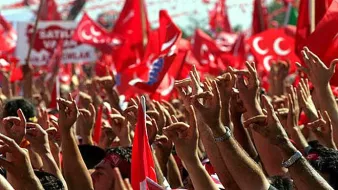 L'image montre une foule rassemblée, brandissant des drapeaux rouges avec des symboles et des motifs typiques. Les participants lèvent les mains, montrant divers gestes de camaraderie ou de soutien. L'ambiance semble festive et engagée, suggérant un événement politique ou une célébration marquée par un fort sentiment nationaliste. Les couleurs dominantes sont le rouge et le blanc, symbolisant souvent l'identité d'un pays.