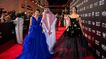 L'image montre trois personnes posant sur un tapis rouge lors d'un événement. À gauche, une femme porte une robe de soirée bleue élégante avec des volants. Au centre, un homme habillé en tenue traditionnelle, comprenant un thobe et un chapeau saoudien. À droite, une femme porte une robe noire ornée de motifs floraux, ajoutant une touche sophistiquée à son look. L'arrière-plan montre un décor stylisé, typique des événements glamour.