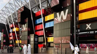 L'image montre une vue d'un bâtiment moderne, probablement un stade ou un centre événementiel, avec une architecture unique. Les murs extérieurs sont composés de containers colorés, alternant des teintes de jaune, rouge et bleu, donnant un aspect dynamique et contemporain. On distingue également des personnes en vêtements traditionnels se tenant près de l'entrée, ainsi que des éléments de sécurité. Le ciel est légèrement nuageux, ajoutant une ambiance particulière à la scène.