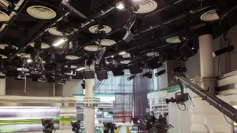 L'image montre un studio de télévision. On peut voir plusieurs caméras montées sur des tréteaux, prêtes à capturer des scènes. Le décor est moderne, avec des surfaces blanches et des accents de couleurs vives, notamment du vert. Des lumières suspendues au plafond éclairent le plateau, et il y a des écrans en arrière-plan, probablement utilisés pour afficher des informations ou des graphiques. L'ambiance semble professionnelle et organisée, typique d'un environnement de diffusion.