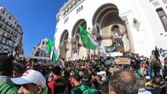 L'image montre une grande foule rassemblée devant un bâtiment avec une architecture traditionnelle. Les manifestants brandissent des drapeaux algériens et des pancartes, exprimant probablement des revendications politiques ou sociales. La scène est en plein air, sous un ciel bleu, et il y a une atmosphère de mobilisation collective, avec des personnes de tous âges vertes et rouges.