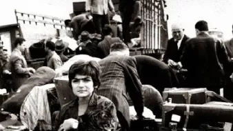 L'image montre une scène de personnes en situation de déplacement. Au premier plan, une jeune fille tient un animal, probablement un chat, dans ses bras, et elle est entourée de nombreux bagages, valises et paquets. En arrière-plan, on aperçoit d'autres personnes, certains sont en train de charger ou de décharger des objets d'un véhicule. L'atmosphère est chargée, et l'image semble capturer un moment de transition ou d'exode, évoquant des sentiments de nostalgie et de désespoir. Les tonalités en noir et blanc ajoutent une dimension dramatique à la scène.