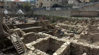L'image montre des fouilles archéologiques en cours dans une zone urbaine. On peut voir des structures en pierre disposées de manière organisée, avec des vestiges de murs et de fondations visibles. Des scaffolds et des passerelles en bois sont installés pour permettre aux archéologues de travailler et d'accéder facilement aux différentes couches de sol. En arrière-plan, des bâtiments modernes sont présents, ce qui témoigne du mélange entre le patrimoine historique et l'urbanisation contemporaine. Des chercheurs et des travailleurs s'activent autour du site, suggérant une recherche active sur l'histoire de la région.