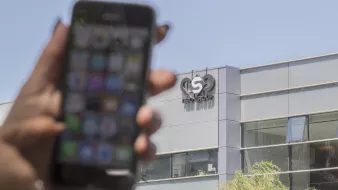 L'image montre un smartphone tenu par une main, flou au premier plan, avec un écran affichant diverses applications. En arrière-plan, on peut voir le bâtiment d'une entreprise, identifié par un logo qui semble être celui d'un groupe nommé "ISN GROUP". Le bâtiment a une façade moderne, probablement en verre. La lumière naturelle éclaire la scène, indiquant qu'il fait jour.