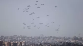 L'image montre un paysage urbain où de nombreux parachutes descendent lentement vers le sol. On peut voir une ville en arrière-plan, avec des bâtiments disposés de manière variable. L'atmosphère semble brumeuse, ce qui donne un aspect à la fois dramatique et intrigant à la scène. Les parachutes indiquent probablement une opération militaire ou humanitaire, créant un contraste avec l'environnement urbain.