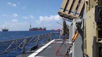 L'image montre un navire en mer, avec des structures de forage pétrolier visibles à l'horizon. On peut apercevoir un système de défense intégrée, probablement dédié à la surveillance ou à la protection, installé sur le pont du navire. Le ciel est clair avec quelques nuages, et la mer est calme, créant une atmosphère maritime paisible. Des chaînes et des équipements de sécurité sont également visibles sur le pont.