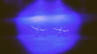 L'image montre deux danseurs sur scène, éclairés par des lumières bleues et violettes. Ils semblent en pleine performance, avec des mouvements expressifs et gracieuses, se trouvant dans un environnement sombre. La lumière crée une atmosphère dramatique, mettant en avant leur gestuelle et l'intensité de leur performance. Le décor est simple, ce qui permet aux danseurs de captiver toute l'attention.