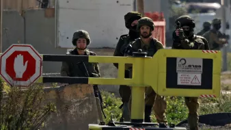 L'image montre des soldats, probablement en situation de contrôle ou de patrouille. Ils portent des uniformes militaires et des équipements de protection. On aperçoit également un panneau de signalisation en forme d'arrêt, qui indique une demande d'arrêt. L'environnement semble être urbain, peut-être dans une zone sensible. Les soldats semblent vigilants et attentifs à leur environnement.