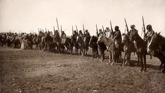 L'image montre une longue ligne de cavaliers en rang, tous montés sur des chevaux. Chaque cavalier est équipé d'une lance ou d'un bâton, et beaucoup portent des vêtements traditionnels. L'arrière-plan semble désertique, avec un sol rugueux et peu de végétation. L'atmosphère de l'image évoque une scène historique ou militaire, mettant en avant des guerriers ou des membres d'une tribu. Les cavaliers adoptent une posture fière et attentive, prêts pour une action ou une procession.