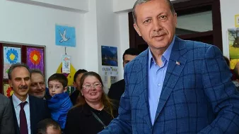 L'image montre un homme d'âge moyen portant un costume bleu. Il est debout, tenant un bulletin dans sa main, et semble être dans une salle de vote ou un endroit similaire. Il y a plusieurs personnes en arrière-plan, dont une femme avec des lunettes, un enfant et d'autres adultes, qui regardent l'homme avec intérêt. L'environnement est chaleureux et coloré, avec des affiches accrochées aux murs.