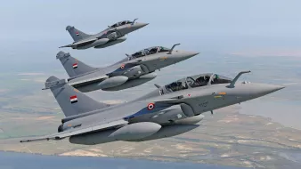 L'image montre un groupe de jets de chasse en plein vol. Trois avions de chasse, probablement des Rafales, sont alignés en formation, avec des couleurs principalement grises. Chaque avion porte des marqueurs et des emblèmes, semblant appartenir à une force aérienne. Le décor en arrière-plan indique un paysage vaste, avec des zones de terre et de l'eau visible, suggérant un vol à basse altitude. L'ensemble dégage une impression de puissance et de précision militaire.