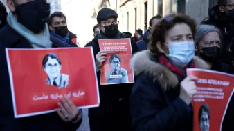 L'image montre un groupe de personnes participant à une manifestation. Elles tiennent des pancartes avec l'image et le nom d'une personne, Farida Adlénah, accompagnées de slogans. Les manifestants portent des masques, probablement en raison de préoccupations sanitaires. L'ambiance semble être solennelle et engagée, avec des visages sérieux. Les arrière-plans montrent des bâtiments qui pourraient avoir une architecture urbaine.