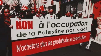 L'image montre une manifestation avec des participants brandissant une grande bannière. Le texte sur la bannière exprime un message de protestation contre l'occupation de la Palestine par Israël, et appelle à ne plus acheter les produits de la colonisation. Les manifestants semblent unis et sont vêtus de vêtements variés, avec des drapeaux en arrière-plan. L'ambiance est sérieuse et engagée, reflétant un message politique fort.