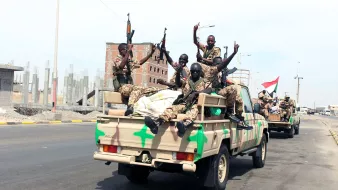 L'image montre un groupe de soldats assis sur le plateau d'un véhicule militaire. Ils portent des uniformes camouflage et tiennent des armes. Les soldats affichent des gestes de victoire en levant un doigt. En arrière-plan, on peut voir un paysage urbain inachevé avec des bâtiments en construction. L'atmosphère semble être celle d'une parade ou d'un mouvement militaire dans une zone urbaine.