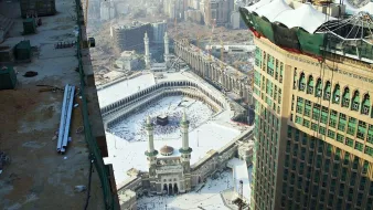 L'image montre une vue aérienne de la Kaaba à La Mecque, en Arabie Saoudite. On peut voir le complexe du Haram qui comprend la mosquée entourant la Kaaba. Il y a de nombreux pèlerins qui s'y trouvent, et l'environnement urbain avec des bâtiments modernes en arrière-plan. On aperçoit également des travaux de construction en cours sur le côté, suggérant une expansion des infrastructures autour du site sacré. Le ciel est légèrement brumeux, ce qui donne une ambiance particulière à la scène.